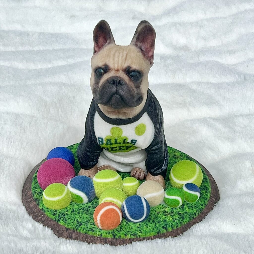 3D printed figurine of a French Bulldog in a black-and-white shirt sitting on grass with multiple colorful tennis balls around it.